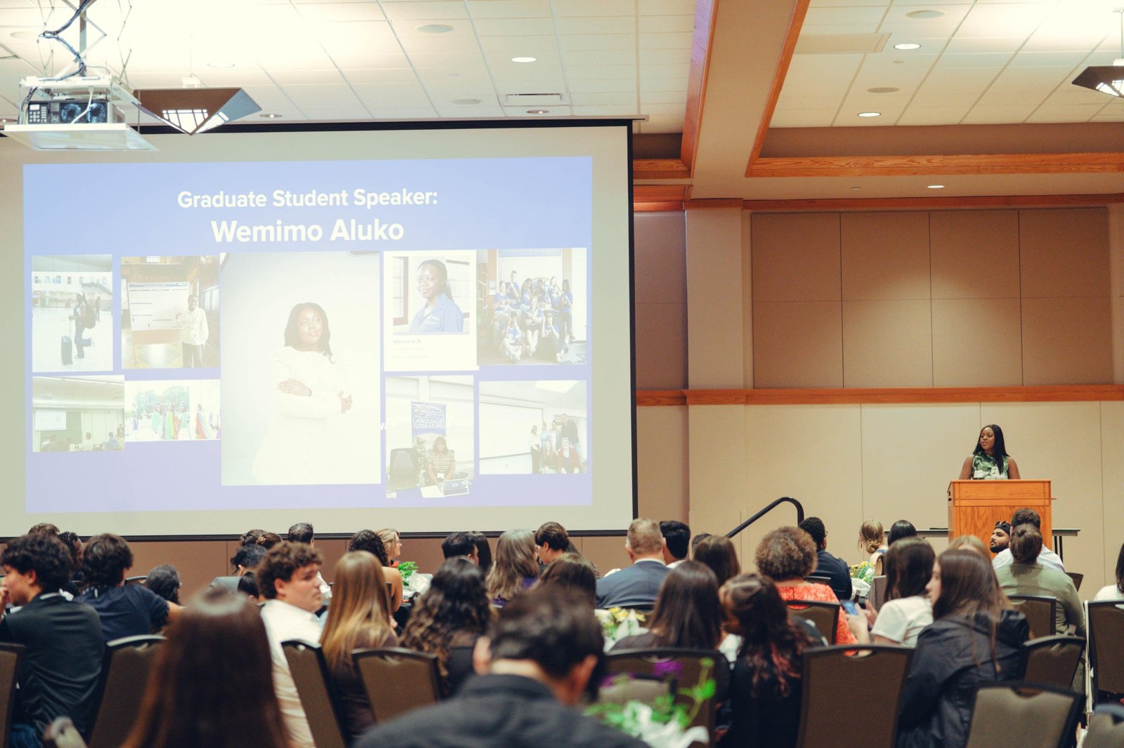 Wemimo speaking at a podium as the graduate student speaker, with her name on the screen
